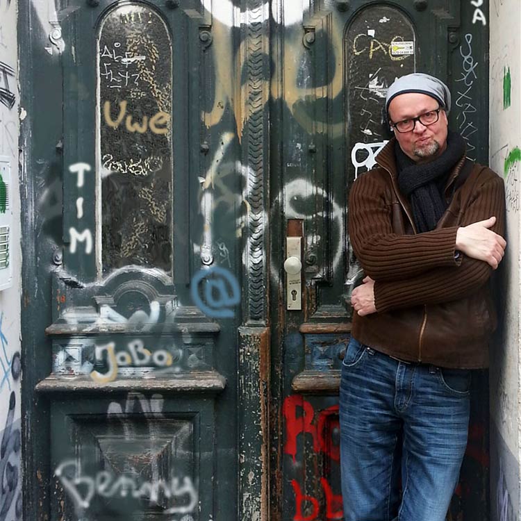 Musiker und Songwriter Mr.Gregg lehnt lässig an der Wand in einem Hauseingang. Die alte, grüne Holztür hinter ihm sowie die darin verbauten schmalen Glasscheiben sind mit vielen, bunten Graffitis besprüht.